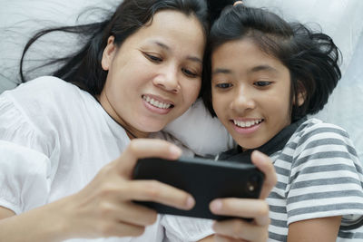Smiling woman with daughter using phone on bed at home
