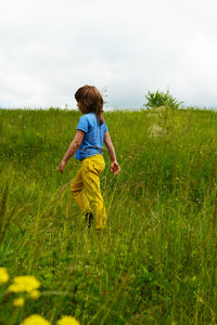 A six-year-old boy in yellow pants and a blue t-shirt with long hair walks in the meadow. 