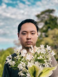 Portrait of young asian man holding bouquet of flowers against cloudy sky and trees.
