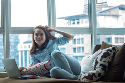 Young woman using phone while sitting on window