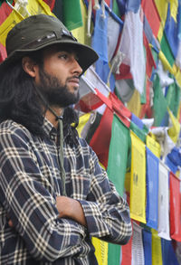 A young guy looking sideways posing with his arms crossed, standing against buddhist prayer flags