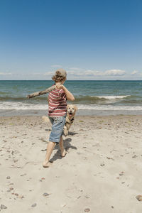 Teenage boy walking by dog on sand at beach against sky