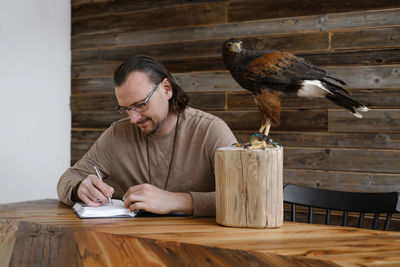 Man is working, writing with wild bird at home by the table. making noted, memories, diary with