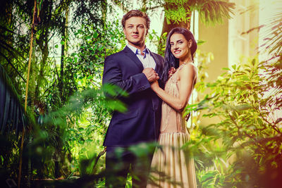 Portrait of smiling bride and groom holding hands while standing amidst plants