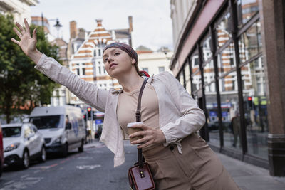 Young woman wearing bandana holding disposable cup waving on footpath