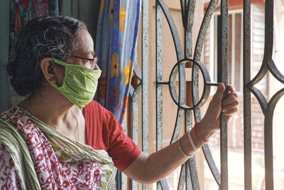 Senior woman looking away from window covered with protective face mask at home