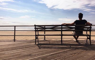 Rear view of man sitting on bench at beach against sky