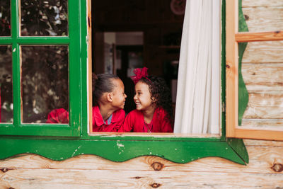Cute black little siblings smiling through open window of wooden cabin