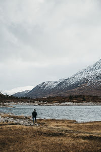 Man standing at lakeshore by mountains against sky during winter