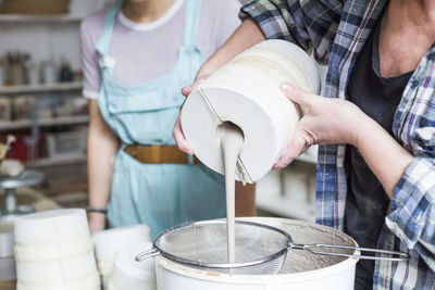 Midsection of mature female potter pouring clay from vase in strainer on bucket at workshop