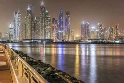 Illuminated modern buildings by river against sky at night