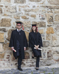 Full length portrait of students with certificate standing against wall