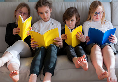 Four children sitting comfortably together on sofa in living room and doing prep work for school