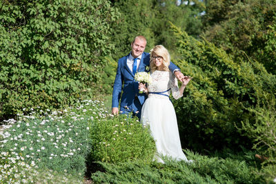 Portrait of smiling bride and groom standing amidst plants at park