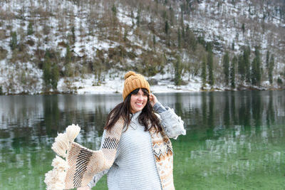 Young woman in winter sweater smiling, standing by a lake.