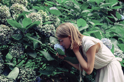 Side view of woman smelling flowers while standing by plants in forest