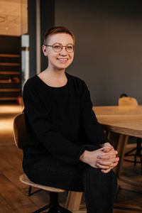 Smiling young woman with short hair in eyeglasses and black sweater sitting at the table in office