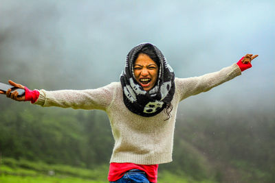 Portrait of cheerful woman with arms outstretched standing outdoors during foggy weather