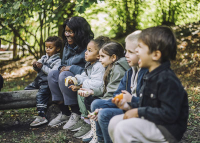 Multiracial boys and girls eating fruits while sitting with female teacher in park