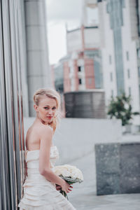 Beautiful bride with bouquet against wall in city