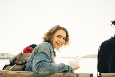 Portrait of cheerful young man holding meal in container while sitting by friend on promenade during sunny day