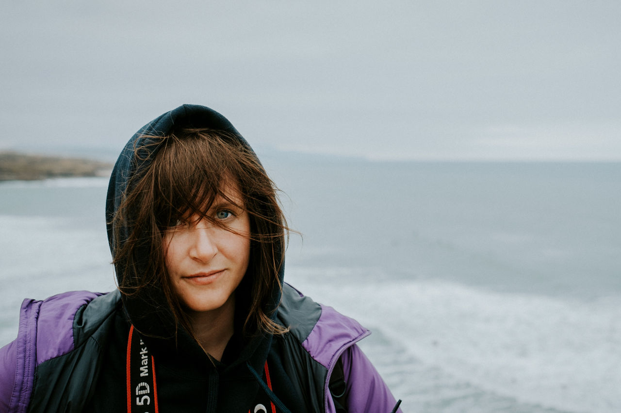 portrait, one person, sea, headshot, water, real people, leisure activity, lifestyles, sky, front view, scenics - nature, beauty in nature, women, nature, non-urban scene, clothing, looking at camera, warm clothing, outdoors, hood - clothing, horizon over water, hairstyle