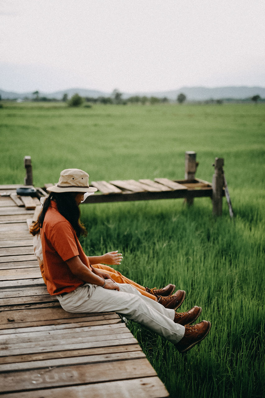 grass, sky, field, one person, land, nature, plant, day, real people, clothing, landscape, hat, green color, wood - material, full length, sitting, leisure activity, lifestyles, relaxation, outdoors