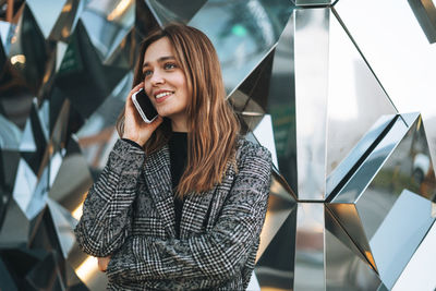 Young smiling woman in coat with coffee cup using mobile phone in evening city street