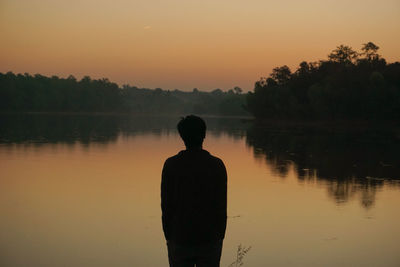 Rear view of man standing by lake against sky during sunset