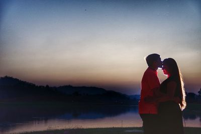 Couple standing by lake against sky during sunset