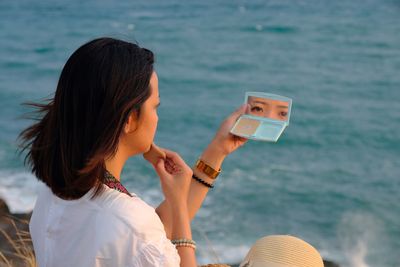 Side view of woman applying make-up sitting by sea