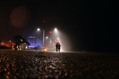 Couple with umbrella walking on street against sky at night during monsoon