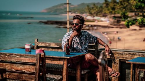 Young man wearing sunglasses while sitting on bench by sea during summer