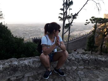 Young woman sitting on retaining wall by mountain against sky