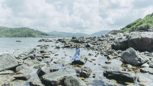 Mid distant view of woman sitting on rocks at beach against sky