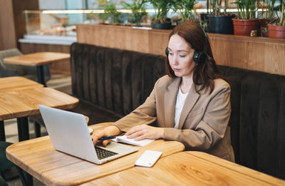 Adult thinking brunette woman forty years in headphones doing notes in daily book with opened laptop