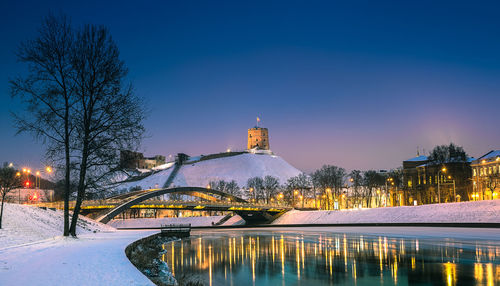 Illuminated buildings by river against sky at dusk