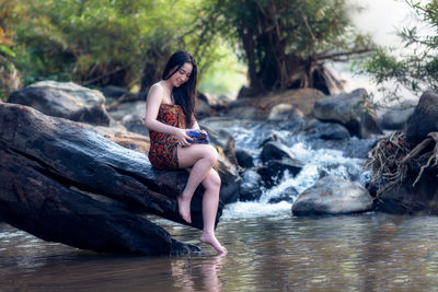 Smiling young woman sitting on rock by stream in forest