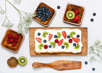 Overhead shot of  strawberry, kiwi, blueberry, raspberry cake and flowers on light gray background.