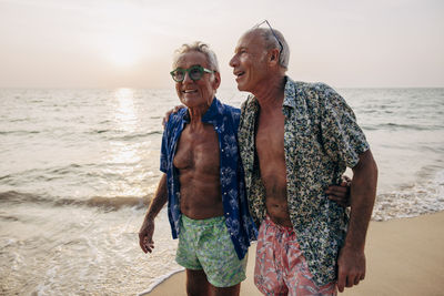 Carefree senior gay couple walking with arms around near shore at beach