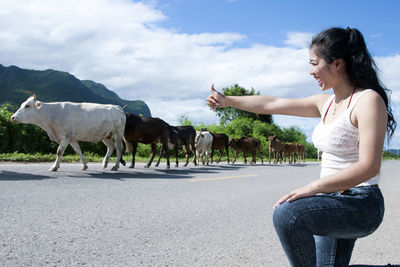 Cheerful woman showing thumbs up to cows walking on road against sky