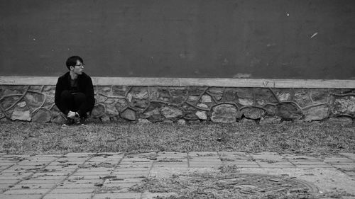 Young man looking away sitting on footpath against wall