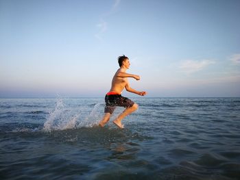 Shirtless boy running in sea against sky