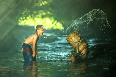 Side view of boy throwing water on man in basket at river