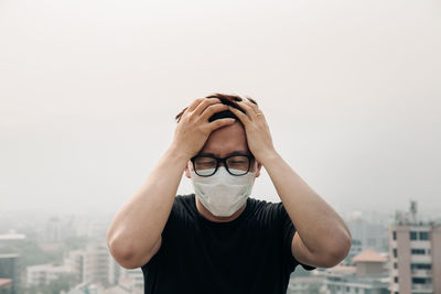 Worried man wearing surgical mask against buildings and sky