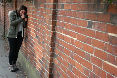 Full length of woman photographing while standing by brick wall