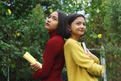 Sisters with back to back standing against trees in park