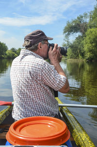 Rear view of senior man photographing while sitting on boat at lake against sky