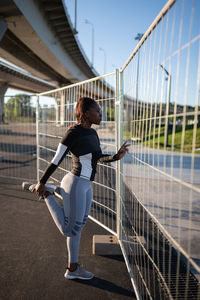 Black sportswoman stretching near fence
