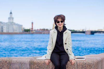 Girl is sitting on granite embankment. in white coat and sunglasses. city